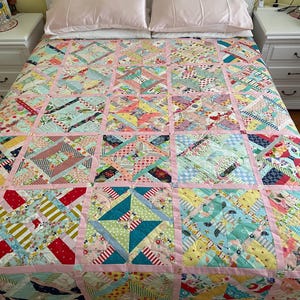 May include: A colorful patchwork quilt with a variety of patterned fabrics in shades of pink, blue, yellow, and green. The quilt is laid out on a bed with pink pillows and a white headboard. The quilt is a mix of geometric shapes.