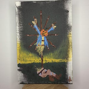 May include: A small canvas painting depicting a scarecrow with a pumpkin head, blue shirt, and straw body. The artwork features a dark background with a green field and a figure lying on the ground. The painting is mounted on a wooden block.