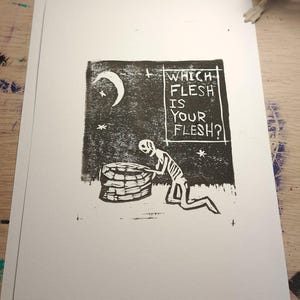 May include: A black and white print on white paper featuring a skeleton kneeling beside a well under a crescent moon and stars. The text "WHICH FLESH IS YOUR FLESH?" is printed in a rectangular box. A small skeleton figurine is in the background.