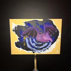 May include: A small, rectangular painting featuring a purple bat with outstretched wings against a dark background. The bat is painted with a cartoonish style, and the painting includes the words "buggy buggy buggies!"