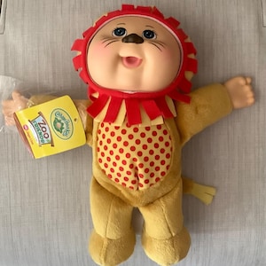 May include: A Cabbage Patch Kids Zoo Friends plush doll dressed as a lion. The doll has a tan body, a red and yellow lion mane hood, and a red polka-dot chest panel. A yellow tag attached to the arm reads Cabbage Patch Kids Zoo Friends Jags Lion.