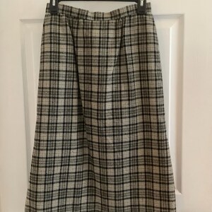 May include: A vintage plaid skirt in a neutral color palette of beige, black, and gray. The skirt is hanging on a black hanger against a white door. The skirt features a classic plaid pattern, with vertical and horizontal lines creating a grid-like design.