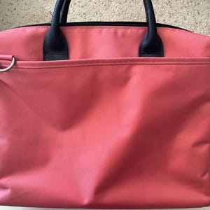 May include: A coral-colored tote bag with black handles and a black zipper. The bag has a front pocket and a silver-colored shoulder strap attachment. The bag is made of a textured material.