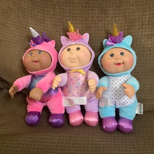 May include: Three plush dolls with round faces and soft bodies, each wearing a hooded unicorn costume. The left doll is pink and purple, the center doll is light purple with a colorful pattern, and the right doll is light blue with white polka dots.