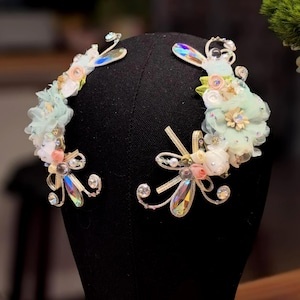 May include: Two decorative headpieces on a black mannequin head. Each headpiece features a floral arrangement with light blue, pink, and white fabric flowers, iridescent crystals, and delicate silver wire accents. The headpieces are designed for special occasions.