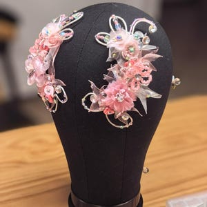 May include: A black mannequin head displays two pink floral hair accessories. The accessories feature fabric flowers, clear crystals, and silver wire accents. The head is on a wooden surface.