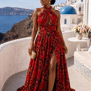 May include: A floor-length red dress featuring a dark patterned print, a halter neckline with a keyhole cutout, and a high side slit. The garment is paired with a small gold clutch, gold earrings, a gold bracelet, and gold strappy sandals.