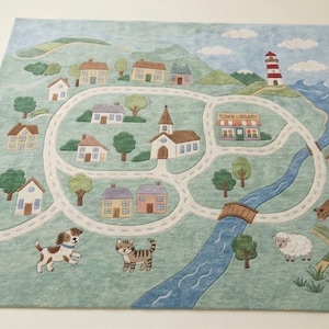 Puede incluir: Una alfombra con temática de pueblo con un fondo azul claro. La alfombra presenta una red de carreteras, casas, una iglesia, una biblioteca, un faro y un río. También se incluyen ilustraciones de un perro, un gato, una oveja y una vaca.