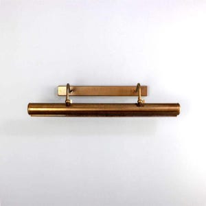 Puede incluir: Una lámpara de latón para cuadros. La lámpara tiene una placa trasera rectangular con dos brazos curvos que sostienen una luz cilíndrica. La luz es de color latón cálido y está montada en una pared blanca.