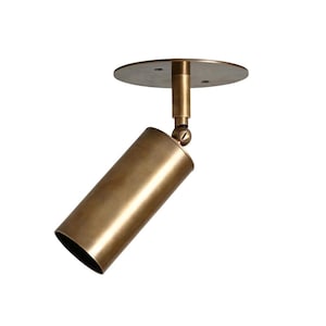May include: A brass-colored, cylindrical spotlight fixture with a circular mounting plate. The light is adjustable with a swivel joint and is designed for ceiling mounting. The fixture has a modern, minimalist design.