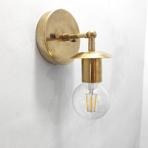 Może przedstawiać: Mosiężny kinkiet z okrągłą płytą tylną i zakrzywionym ramieniem podtrzymującym skierowaną w dół oprawę oświetleniową. Oprawa ma cylindryczną podstawę i mały, okrągły klosz, w którym znajduje się przezroczysta, okrągła żarówka.