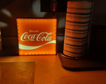May include: Illuminated Coca-Cola sign with the text "Bevete Coca-Cola" in white on an orange background. A roll of paper towels is to the right. The scene is lit with warm, orange light.