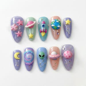 May include: A set of ten long, oval-shaped artificial nails with a space theme. Each nail is painted in a different pastel color and decorated with 3D space-themed embellishments, including stars, planets, rockets, and aliens. The nails have a glittery finish.