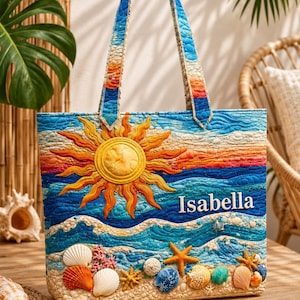 May include: A quilted tote bag with a beach theme, featuring a sun, waves, and seashells. The name "Isabella" is embroidered on the front. The bag has blue, orange, and white colours. The handles are blue and orange.