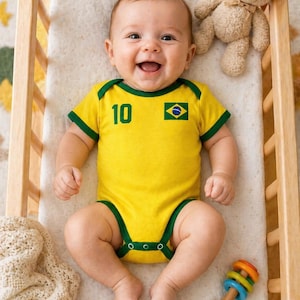 May include: A smiling baby wearing a yellow bodysuit with green trim and the number 10. The bodysuit features the Brazilian flag. A teddy bear and a colorful rattle are also in the wooden crib.