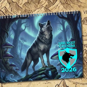 May include: A spiral-bound calendar featuring a detailed illustration of a gray wolf howling at the sky in a forest setting. The calendar is labeled "Dire Wolf Calendar 2026" with a wolf emblem. The background includes trees, mushrooms, and an aurora borealis.