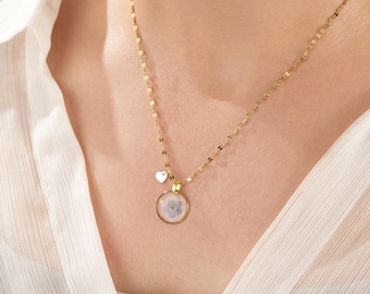 Puede incluir: Un collar dorado con un colgante redondo que contiene una pequeña flor azul. Un pequeño dije de corazón cuelga cerca del colgante. El collar se usa sobre una blusa blanca.