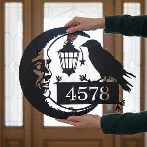 Pode incluir: Placa de endereço de metal preto com uma lua crescente com um rosto, um corvo empoleirado e uma lanterna suspensa. A placa exibe o número 4578. O design é segurado por mãos contra um fundo desfocado.