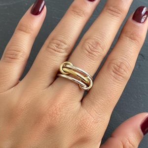 May include: A close-up of a two-tone ring with a gold and silver design. The ring features a series of interlocking loops, with a gold loop in the center and silver loops on either side. The ring is on a hand with burgundy nail polish.