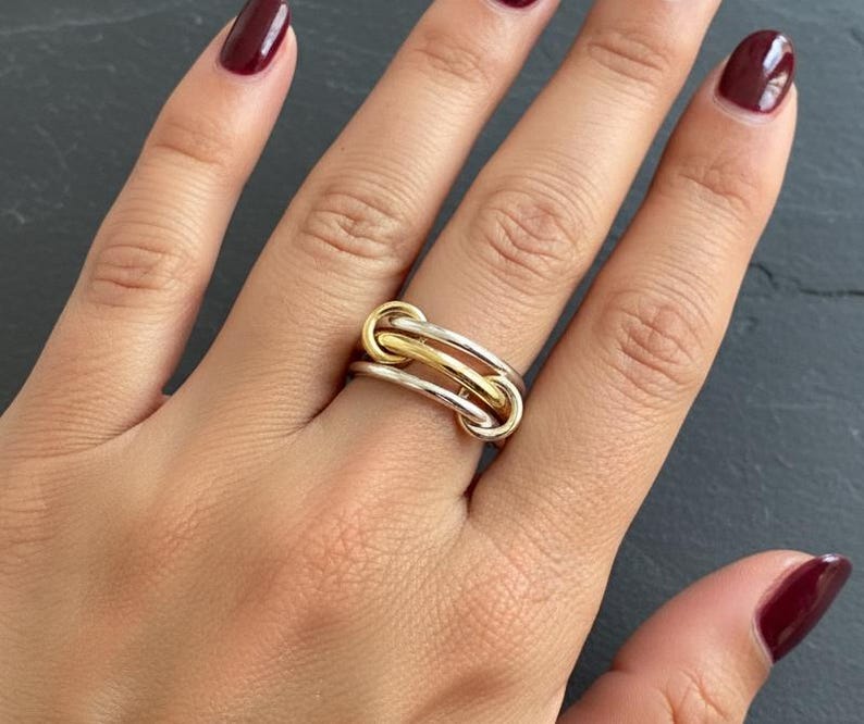 May include: A close-up of a two-tone ring with a gold and silver design. The ring features a series of interlocking loops, with a gold loop in the center and silver loops on either side. The ring is on a hand with burgundy nail polish.