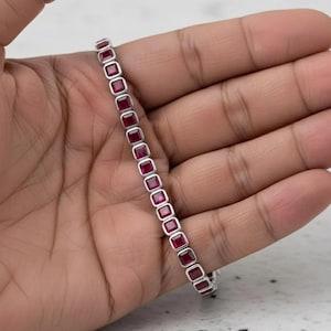 May include: A silver bracelet featuring a line of square-cut, deep red gemstones. The gemstones are set in a silver band, creating a continuous pattern. The bracelet is shown against a neutral background.