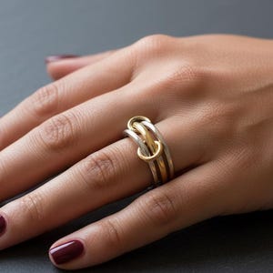 May include: A silver and gold interlocking ring with three connected bands. The ring is worn on a hand with dark red nail polish. The ring's design features a gold band intertwined with two silver bands.