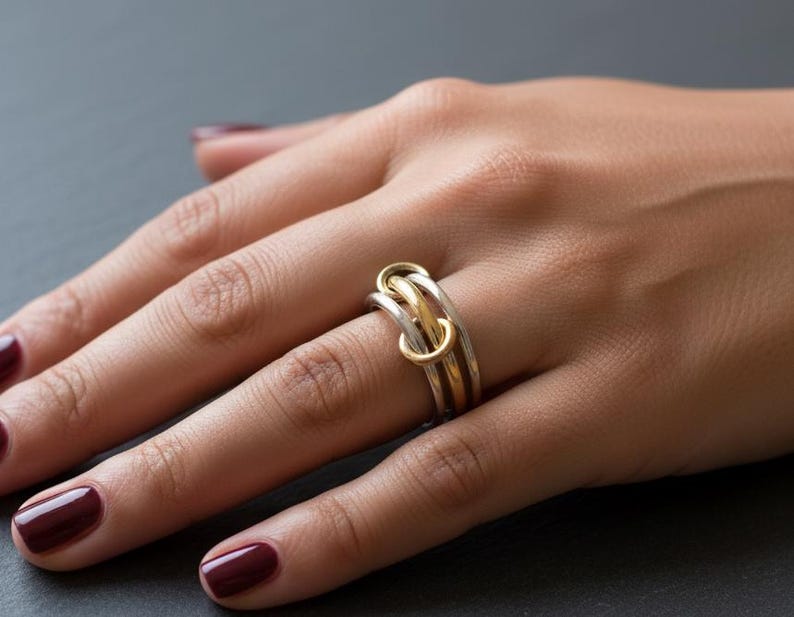 May include: A silver and gold interlocking ring with three connected bands. The ring is worn on a hand with dark red nail polish. The ring's design features a gold band intertwined with two silver bands.
