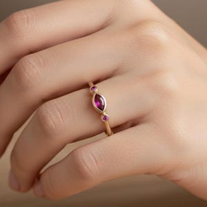Könnte beinhalten: Ein goldener Ring mit einem markisenförmigen, tiefroten Edelstein, flankiert von zwei kleineren, runden, rosa Edelsteinen. Der Ring ist auf einem schlanken Goldband gefasst und zeigt ein zartes und elegantes Design. Schmuck.
