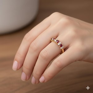 以下が含まれることがあります： 赤色の宝石と小さな白い宝石が交互に並んだゴールドリング。指にはめられており、ジュエリーのデザインと色のコントラストが際立っています。リングのバンドは細く、磨かれたゴールドです。