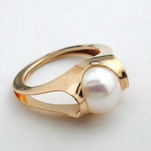 May include: A gold-colored ring featuring a large, round, white pearl. The ring has a modern design with a split band and a setting that cradles the pearl. The ring is set against a white background.
