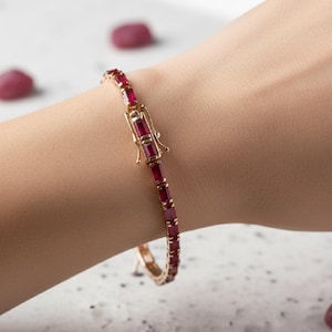 Puede incluir: Una pulsera dorada con piedras preciosas rectangulares de color rojo intenso. La pulsera se cierra con un cierre. La joya se muestra en una muñeca, destacando el diseño y el color.