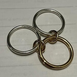 May include: Three interlocking rings, two silver and one gold, create a unique design. The rings are smooth and circular, with a polished finish. The gold ring adds a contrasting element to the silver rings.
