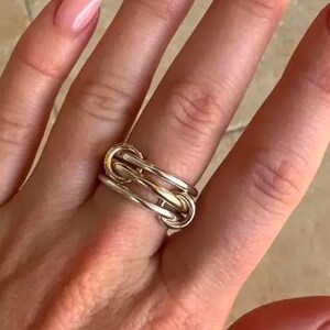 May include: A two-tone ring with a linked design. The ring features a combination of silver and gold tones, creating a modern and stylish look. The ring is worn on a finger, showcasing its intricate design and metallic finish.