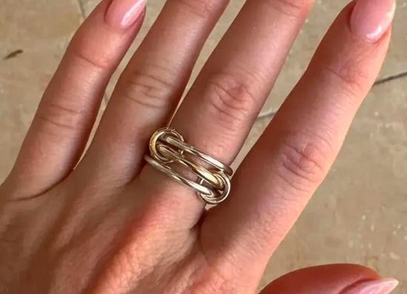 May include: A two-tone ring with a linked design. The ring features a combination of silver and gold tones, creating a modern and stylish look. The ring is worn on a finger, showcasing its intricate design and metallic finish.