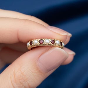 Op de afbeelding: Een gouden ring met afwisselend ronde parels en rode edelstenen. De ring wordt tussen vingers gehouden tegen een blauwe achtergrond. De edelstenen zijn gelijkmatig verdeeld over de band, wat een klassiek en elegant ontwerp creëert.