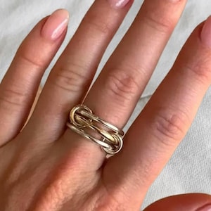 Può includere: Un anello dorato e argentato con un design a nodo. L'anello è indossato su un dito, mostrando il suo intricato motivo intrecciato. Il design dell'anello presenta una combinazione di metallo lucido e strutturato.