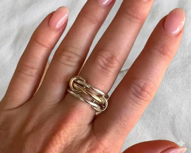May include: A gold and silver-toned ring with a knot design. The ring is worn on a finger, showcasing its intricate interwoven pattern. The ring's design features a combination of polished and textured metal.