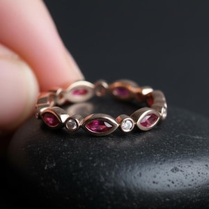Peut inclure: Une bague d'éternité en or rose ornée d'alternances de rubis taillés en marquise et de diamants ronds. La bague est tenue par un doigt sur un fond sombre. Les pierres précieuses sont d'un rouge vif.
