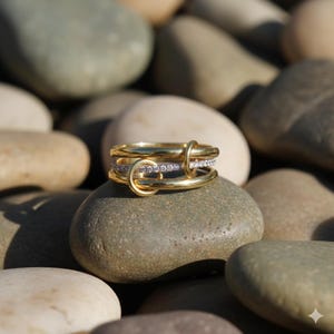 Pode incluir: Um anel dourado com três faixas, uma delas com pequenas pedras transparentes. O anel tem um design único com um elemento em forma de laço que conecta as faixas. O anel é exibido em uma pedra lisa e cinza.