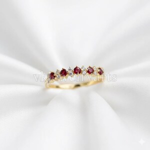 Peut inclure: Une bague en or ornée d'une rangée alternée de pierres précieuses rouges et de petites pierres blanches. La bague est posée sur un fond blanc. Il s'agit d'un bijou.