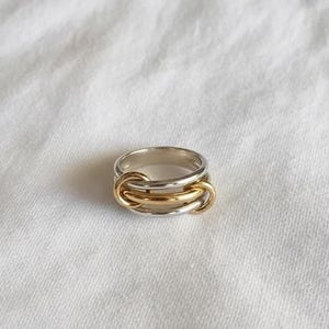 May include: A silver and gold ring with a unique interlocking design. The ring features a silver band with three gold-colored interlocking loops in the center. The ring is set against a white, textured background.