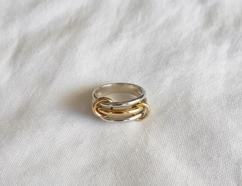May include: A silver and gold ring with a unique interlocking design. The ring features a silver band with three gold-colored interlocking loops in the center. The ring is set against a white, textured background.