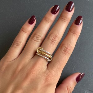 May include: A multi-tone ring with a linked design. The ring features silver and gold tones, with a polished finish. The ring is worn on a finger with burgundy nail polish. The background is a dark gray surface.