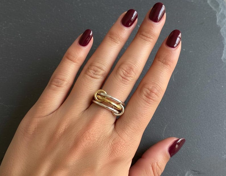May include: A multi-tone ring with a linked design. The ring features silver and gold tones, with a polished finish. The ring is worn on a finger with burgundy nail polish. The background is a dark gray surface.