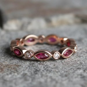 Op de afbeelding: Roségouden bandring met afwisselend marquise-geslepen robijn edelstenen en ronde diamanten. De ring ligt op een getextureerd grijs oppervlak. De edelstenen zijn helderrood.