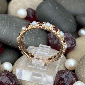 Könnte beinhalten: Ein goldener Ring mit abwechselnd roten Edelsteinen und weißen Perlen. Der Ring wird auf einem transparenten Ständer präsentiert, umgeben von Steinen und Perlen. Das Band des Rings ist aufwendig gestaltet.