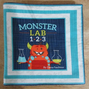 May include: A blue and white fabric book cover with a monster illustration and the title "Monster Lab 1.2.3" by Dana Saulnier.