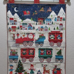 May include: A white fabric advent calendar with a Christmas train theme. The calendar has 24 numbered pockets, each with a different festive image. The train is decorated with red, green, and blue colors. The calendar is hung on a green rod with red and gold accents.
