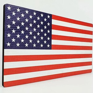 May include: Wooden American flag with a distressed finish. The flag features the traditional red and white stripes and a blue field with white stars. The wood grain is visible, adding texture to the patriotic design.