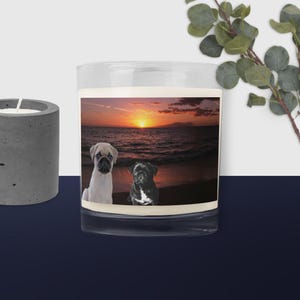 May include: A clear glass candle holder with a sunset scene featuring two pugs on a beach. One pug is light beige, the other is black and white. The candle holder is next to a grey concrete candle holder.